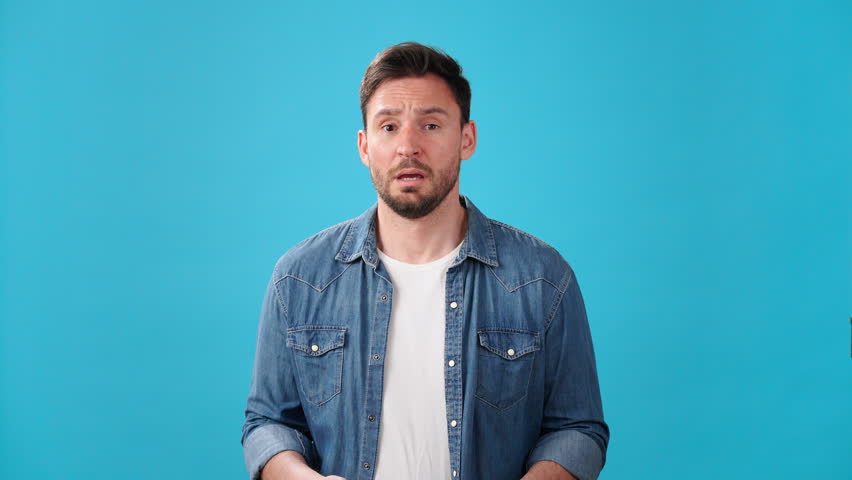Nervous man takes deep breath and shows palms trying to calm down. Worried young person wipes forehead showing excitement on blue background | Shutterstock HD Video #1108937867