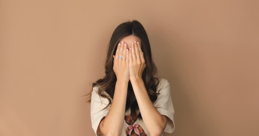 Positive smiling young woman peeking through fingers with curious nosy expression, spying secret information standing on beige background. She open face and laughing. Girl look happy, wow emotions.