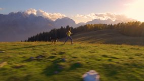 AERIAL: Woman jogging with her dog on top of alpine meadow hill in golden sunset. She and her cute doggo run fast along a grassy ridge with a stunning mountain backdrop on a picturesque autumn day.