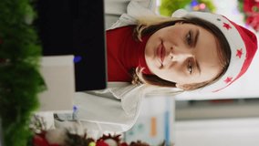 Vertical video Focused woman in Christmas ornate office looking at computer screen, and drinking coffee. Diligent employee doing research in diverse xmas adorn workplace during festive holiday season