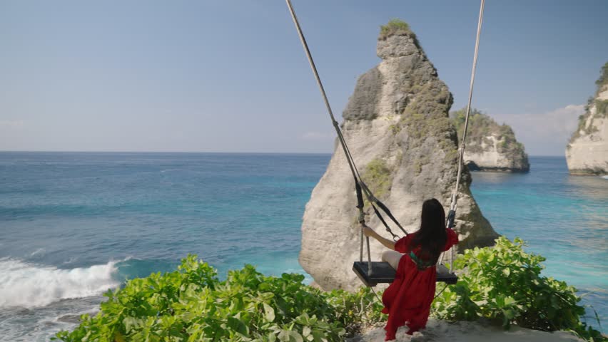Nusa penida island, Bali, indonesia. 4K video Young woman red dress Play on the swings at Diamond beach, beutiful diamond beach,Nusa Penida island. Popular travel destination on Bali, indonesia.