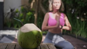 Mid adult woman sitting at namaste position, green fresh coconut is ready for drinking, outdoors exercises