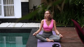 Mid adult woman sitting in lotus position on yoga mat near the swimming pool at daytime