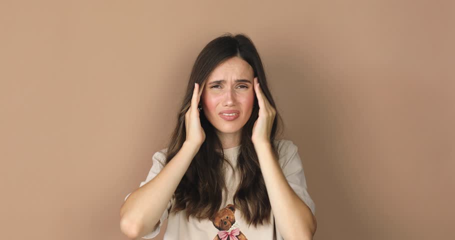 Attractive woman close up with strong headache. Beautiful young brunette touching her temples feeling stress or pain. Stress or headache concept. Negative human emotion isolated on beige background.