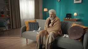 Zoom in video of an eldery, retired man, senior citizen sitting on a couch, sofa wrapped in blanket, coughing as if he has a flu or cold. He used lots of tissues for his runny nose.