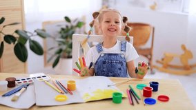 a little child girl happily makes prints in an album with her hands with a dirty face and smiles, a happy child draws a picture with gouache fingers at home at the table - Powered by Shutterstock - Get 15% off with code: PIKWIZARD15