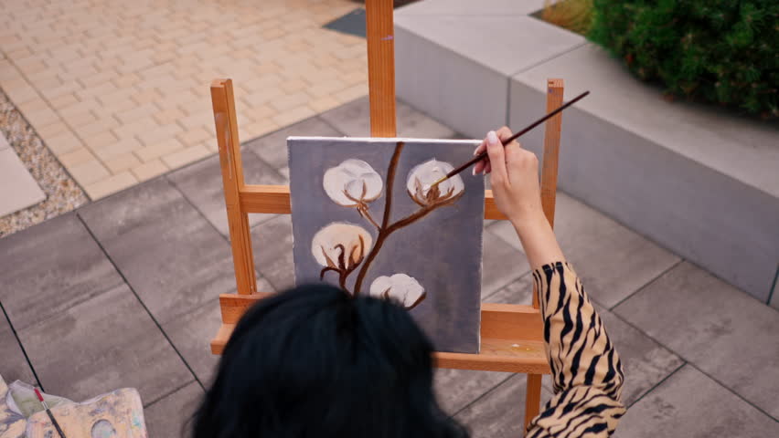 A young girl artist paints picture with a brush on a canvas that stands on an easel view from behind the back