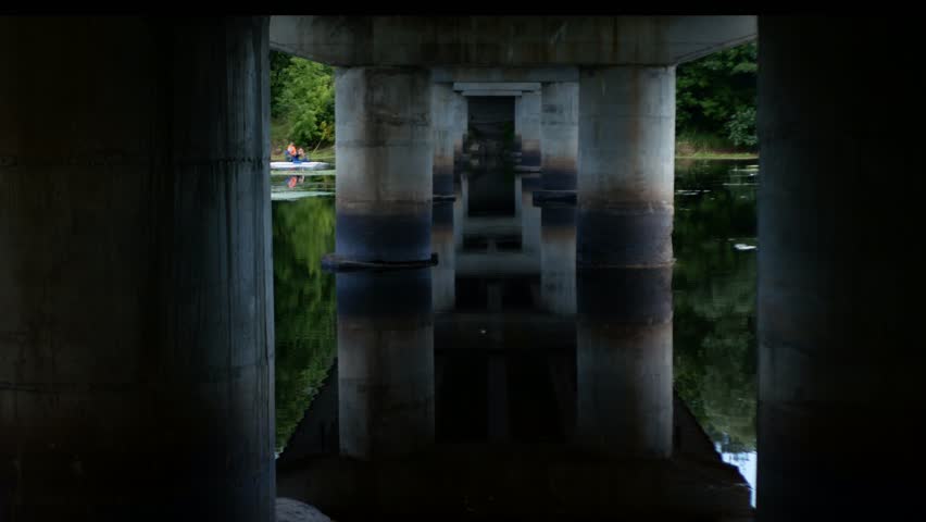 People in kayaks pass under a railway bridge with massive supports. On the massive pillars it is noticeable that the water level has dropped.