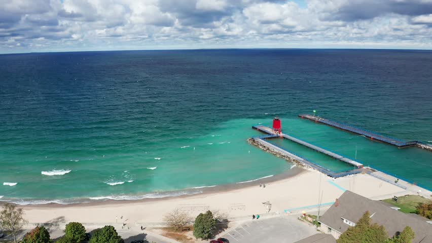 Drone footage circling a pier with a lighthouse and a channel leading to lake Michigan. The sky is cloudy and it is day. The water is deep and blue.