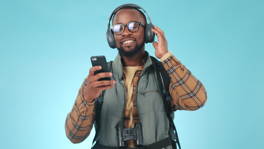 Dancing, smile and black man with a smartphone, headphones and streaming music on a blue studio background. African person, guy or model with headset, cellphone and digital app with energy and moving | Shutterstock HD Video #1108982781