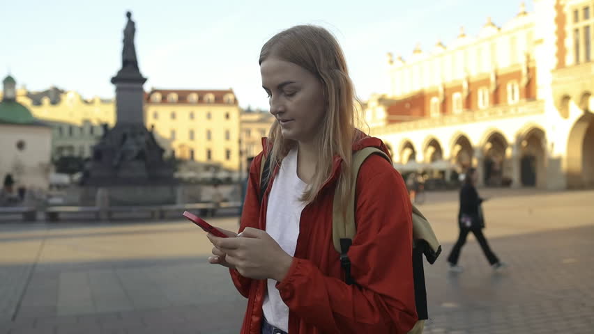 St. Marys Basilica. Positive young Caucasian woman with yellow backpack using navigation app on phone and walking in old city while travelling alone on Market Square in Krakow. Traveling Europe. High