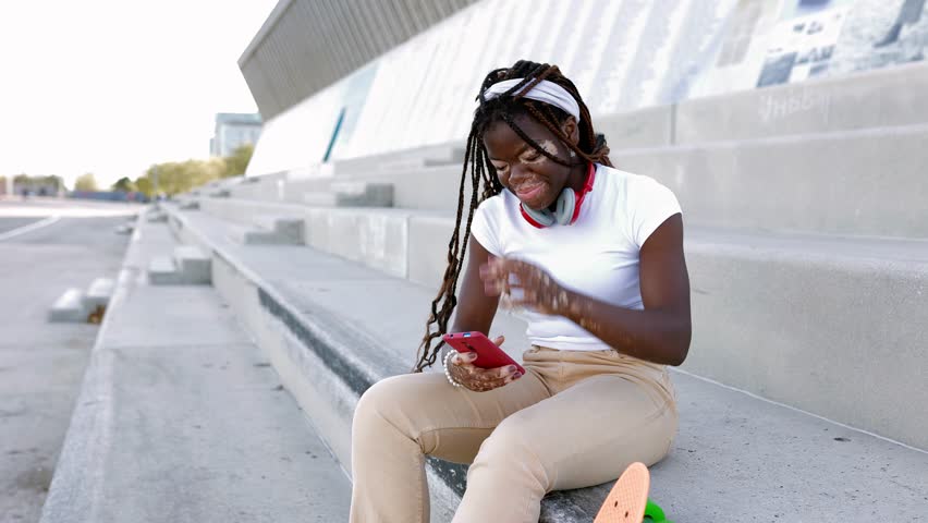 Young african teenage girl with vitiligo having fun using mobile phone sitting outdoors. Youth culture and technology concept. Copy space for text. | Shutterstock HD Video #1108984313