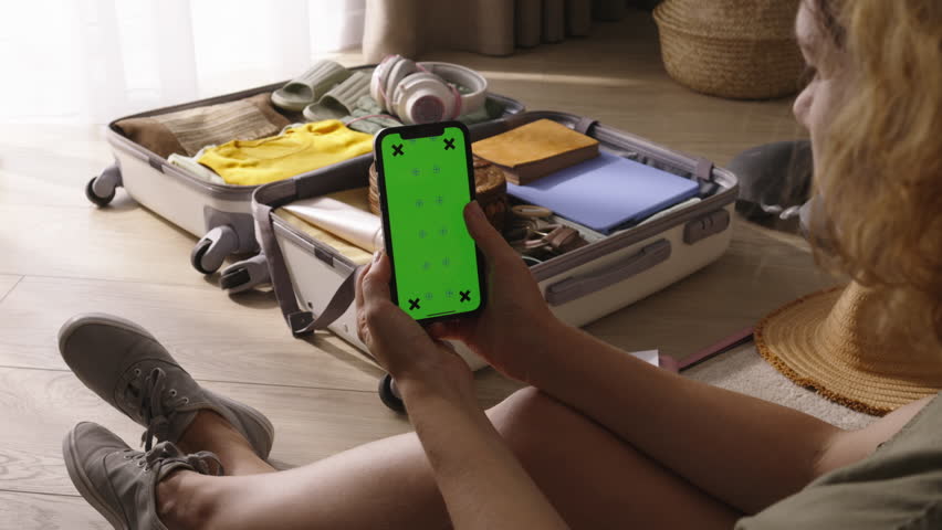 Woman scrolls browses smartphone with green screen. Chroma key on smartphone screen woman traveler against of collected luggage in suitcase looks buys cheapest plane tickets for long journey vacation