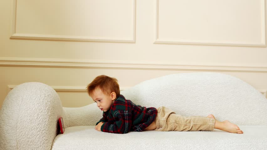 A blond boy lies on his stomach on the sofa and watches cartoons on the phone. the child looks at the phone screen | Shutterstock HD Video #1108997855