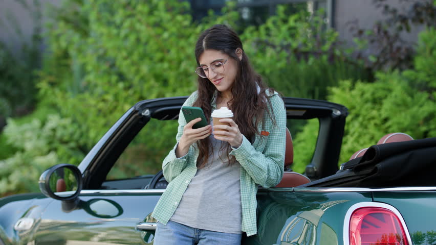 Woman Browsing Smartphone Drinking Coffee Standing Outside near Car. Female scrolling her cell phone near her convertible. | Shutterstock HD Video #1108998335