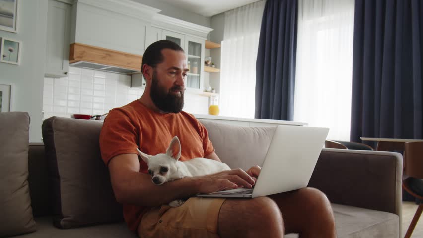 Adult man works with one hand on a laptop and at the same time strokes his little cute dog. An freelancer guy work from home online with his lovely pet