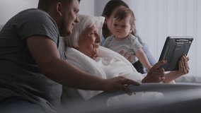 Hospital ward. Elderly woman lying in bed spends time with family. Loving relatives use digital tablet, support grandmother recovering after successful surgery. Modern medical facility. Slow motion.