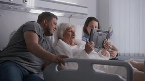 Hospital ward. Loving family spends time together, talks by video call using tablet computer. Relatives support old grandmother recovering after successful surgery. Modern medical facility or clinic.