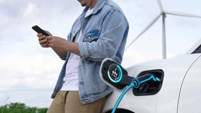 Asian man use phone while electric car recharge energy, showing digital battery status hologram from EV charger from charging station at wind turbine farm. Alternative clean energy utilization.Peruse - Powered by Shutterstock - Get 15% off with code: PIKWIZARD15