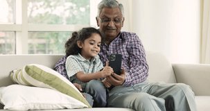 Loving multi-generational family, pretty preschooler 5s great-grandson and older 70s great-grandfather spend time on internet hold mobile phone, use new app, discuss video game, rest on sofa at home