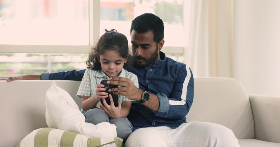 Loving Indian dad teach little adorable son to using modern smartphone, learn new mobile application, play on-line games resting on sofa at home. Younger generation and wireless tech, internet overuse | Shutterstock HD Video #1109028247