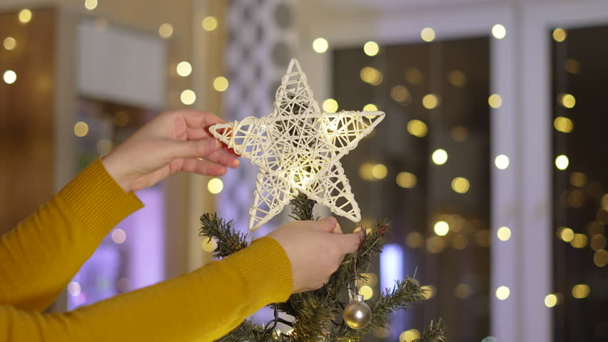 Unrecognizable woman touches main decoration of Christmas tree. Person carefully adjusts symbol of birth of Jesus Christ, star of Bethlehem. Eco friendly toy on top of traditional tree in living room.