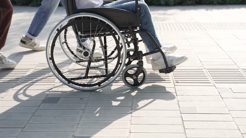 Young friends pushing wheelchair of disabled man, having fun together walking in the city park