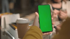 View from shoulder of caucasian woman doing swipe up gesture on chroma key of modern smartphone. Female relaxing in a restaurant and using cell phone.
