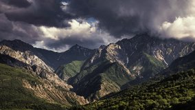 Timelapse mountains. Huge rocky mountain range with green foot. Greater Chimgan Kyzylzhar. Heavy thunderclouds swirl over tops of mountains illuminated by rays of sun. Cinematic video for background