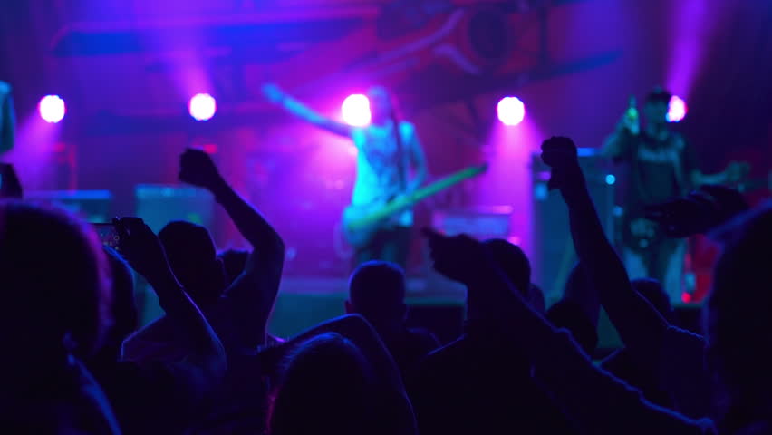 Rock Band with Guitarists and Drummer Performing at a Concert in a Night Club. Front Row Crowd is Partying. Silhouettes of Fans Raise and wave Hands in Front of Bright Colorful Strobing Light on Stage Royalty-Free Stock Footage #1109048351