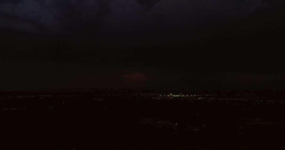 Cinematic aerial of dark storm clouds with dramatic lightning bolts flashing in the dark, thick clouds at night above a small town as the city light twinkle below in this stormy establishing shot.