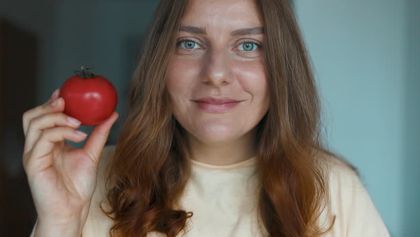 Smiling young woman showing tomato and lookin at camera, as she prepares dinner. Girl licks her lips. High quality FullHD footage