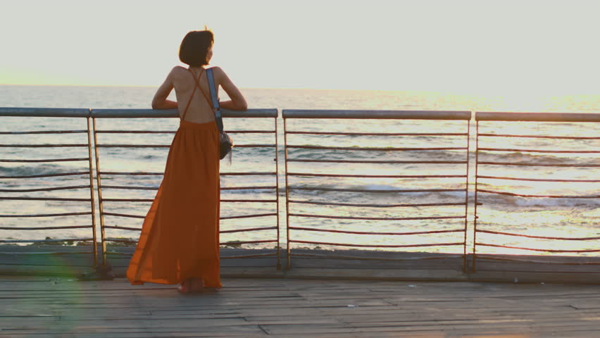 A woman enjoys the sunset while traveling. Elegant happy woman in a dress looks at the ocean.