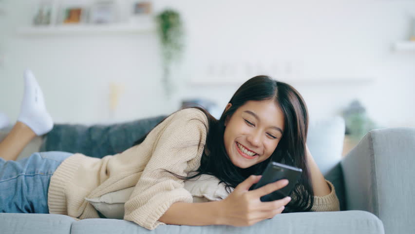 Happy young asian woman relax on comfortable couch at home texting messaging on smartphone, smiling girl use cellphone, browse wireless internet on gadget, shopping online from home | Shutterstock HD Video #1109056085