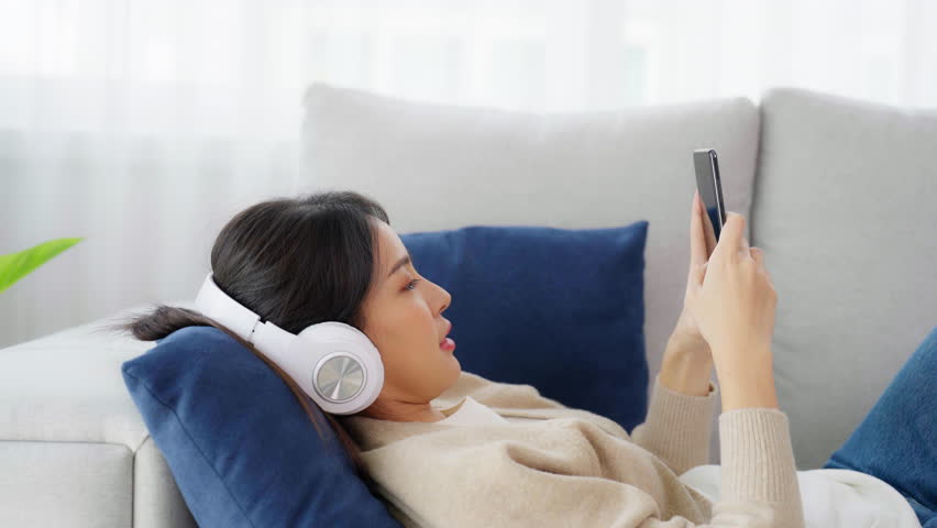 Happy young asian woman wearing headset and relax on comfortable couch at home texting messaging on smartphone, smiling girl use cell phone video call | Shutterstock HD Video #1109056087