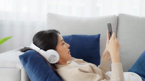 Happy young asian woman wearing headset and relax on comfortable couch at home texting messaging on smartphone, smiling girl use cell phone video call