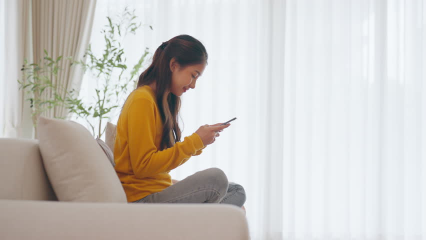 Happy young asian woman relax on comfortable couch at home, typing chat message on smartphone, smiling girl use cell phone chatting, searching information on browser wireless internet, online shopping | Shutterstock HD Video #1109056097