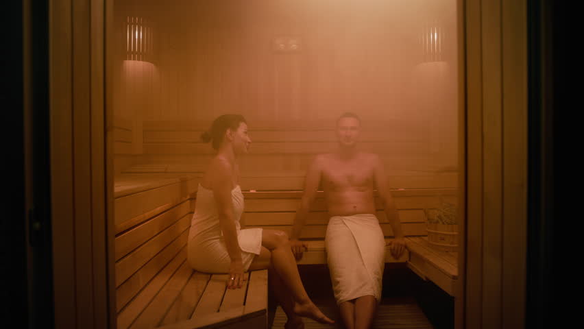 A middle-aged man and a woman relax in a Finnish sauna. They are having a conversation. Both of them are wrapped in towels. They are sitting on the wooden benches. The sauna is dimly lit and steamy. Royalty-Free Stock Footage #1109059527