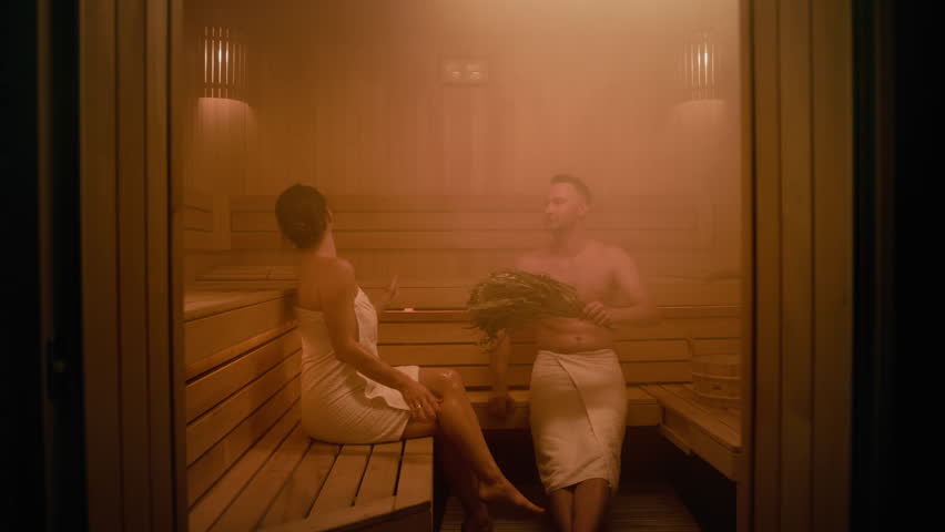 A woman and a man are sitting in a Finnish sauna. They are both covered with towels. The man waves a whisk over himself and puts it in a bucket. The woman says something to the man and he replies. Royalty-Free Stock Footage #1109059539
