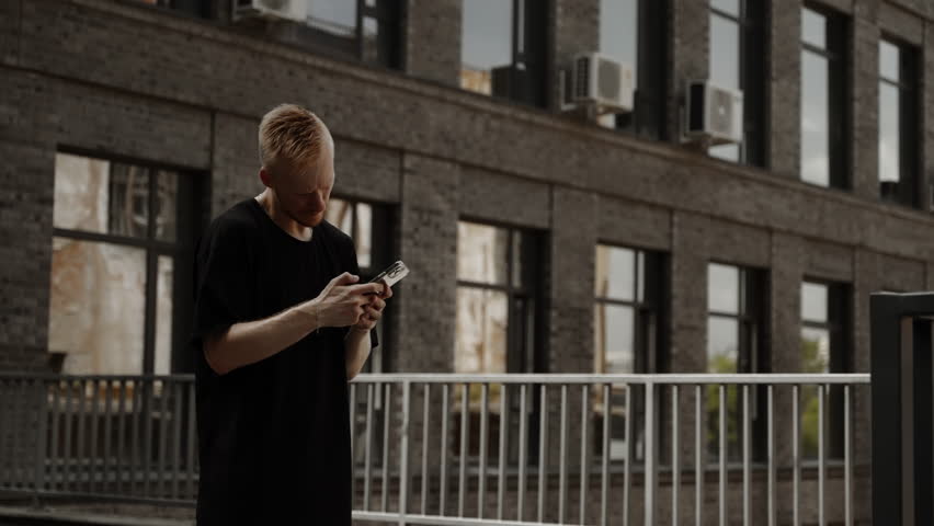 A man chooses music on a smartphone. A young man in a black T-shirt stands in front of a building. Young man in black t-shirt with mobile phone in the city | Shutterstock HD Video #1109059579