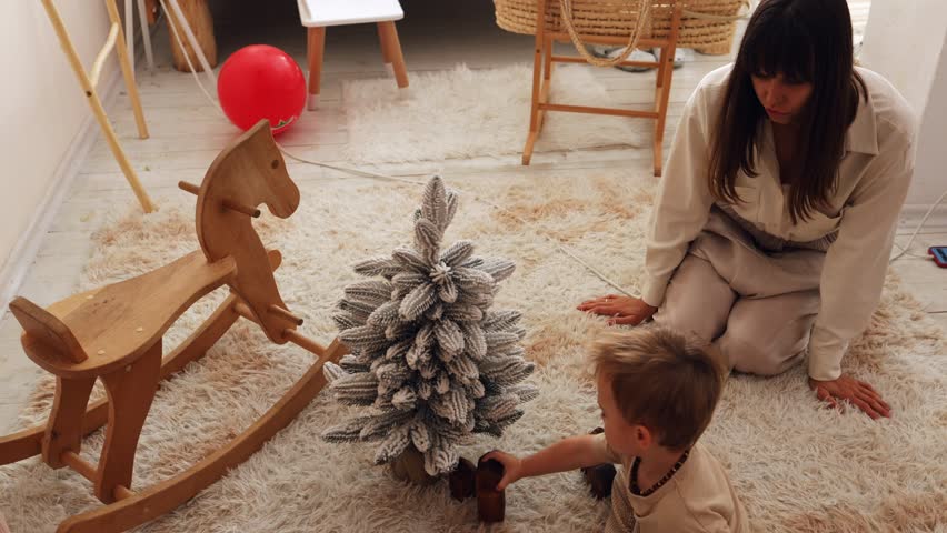 mother plays with her little son near a small artificial Christmas tree at home near a wooden horse. the child plays with wooden toys near the Christmas tree