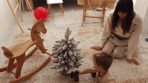 mother plays with her little son near a small artificial Christmas tree at home near a wooden horse. the child plays with wooden toys near the Christmas tree Adlı Stok Video