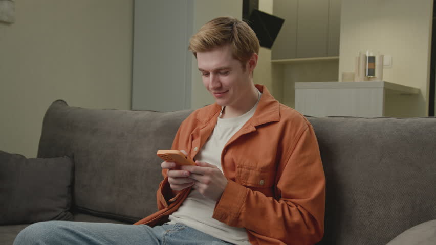 Man Browsing Cell Phone. White person surfing the net using smartphone. Smiling young adult holding phone sitting on couch. | Shutterstock HD Video #1109069893