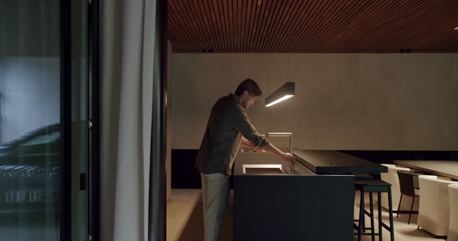 Young man goes to kitchen island with sink, take glass and fill drink water from faucet at modern apartment with stylish furniture and interior.