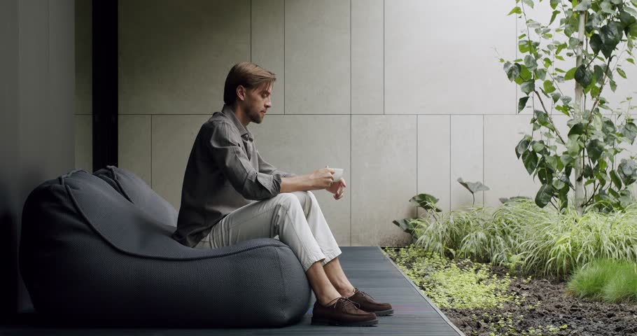 Young man in stylish clothes sitting on comfortable bean bag armchair on terrace, resting on nature and drink coffee enjoying morning at summer day outdoors. Male relax on frameless chair outside. Royalty-Free Stock Footage #1109072343