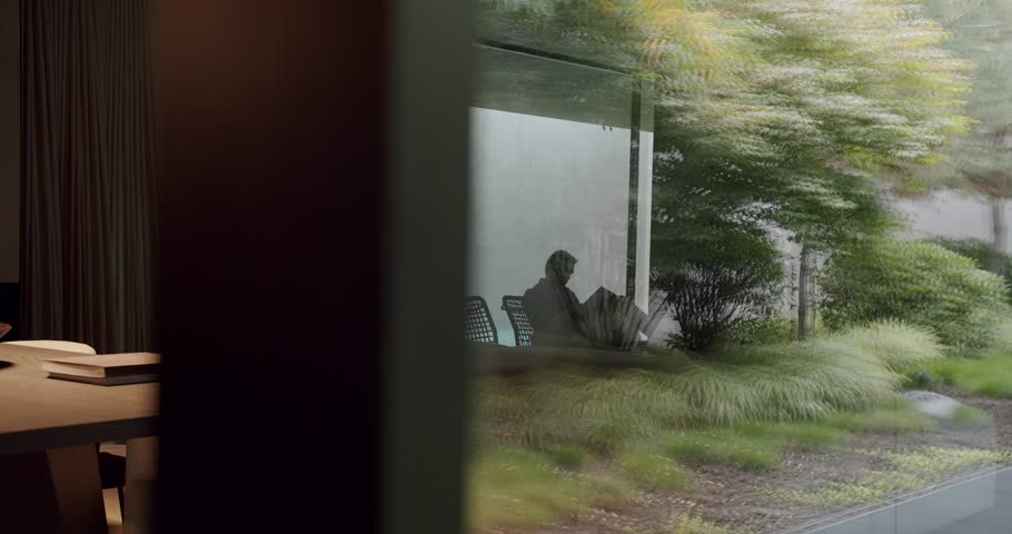 Man sitting in comfort chair and read book at luxury apartment behind glass wall with reflection of garden. Male resting with magazine inside modern cottage house with glass door or window Royalty-Free Stock Footage #1109072369