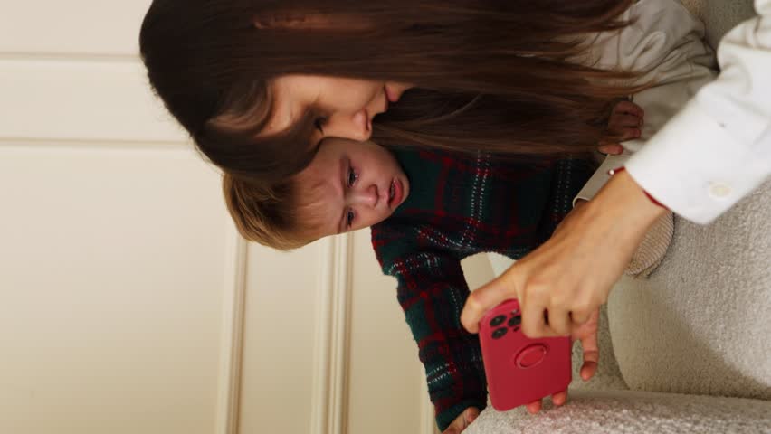 Mother soothes her little son who is crying on the couch. the boy wipes his tears with his hands and turns to the red phone where his mother is showing cartoons. vertical video | Shutterstock HD Video #1109073343