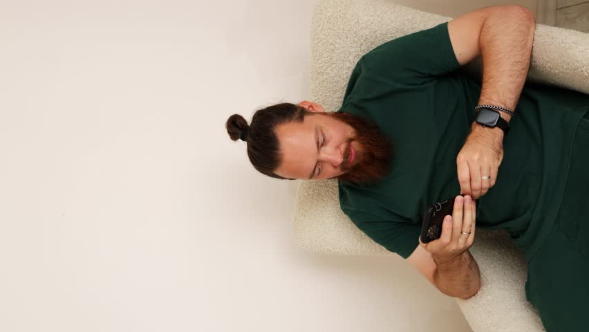 A stylish guy in a green t-shirt with hair tied in a bun and a red beard is sitting in a chair on the phone and looking at the camera and smiling. vertical video | Shutterstock HD Video #1109073351