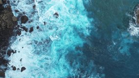 This aerial drone video captures the azure turquoise ocean, the towering cliffs, rugged stone beach, and the relentless crashing of waves against the shore in Bali