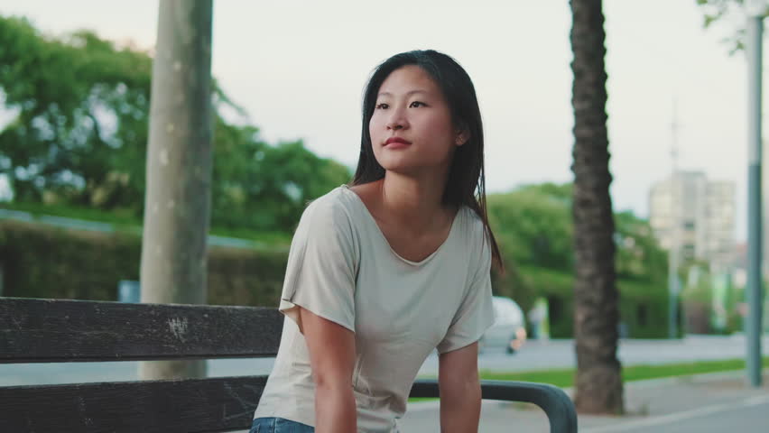 Beautiful happy brunette girl dressed in casual clothes, sits on bench outside, looks around